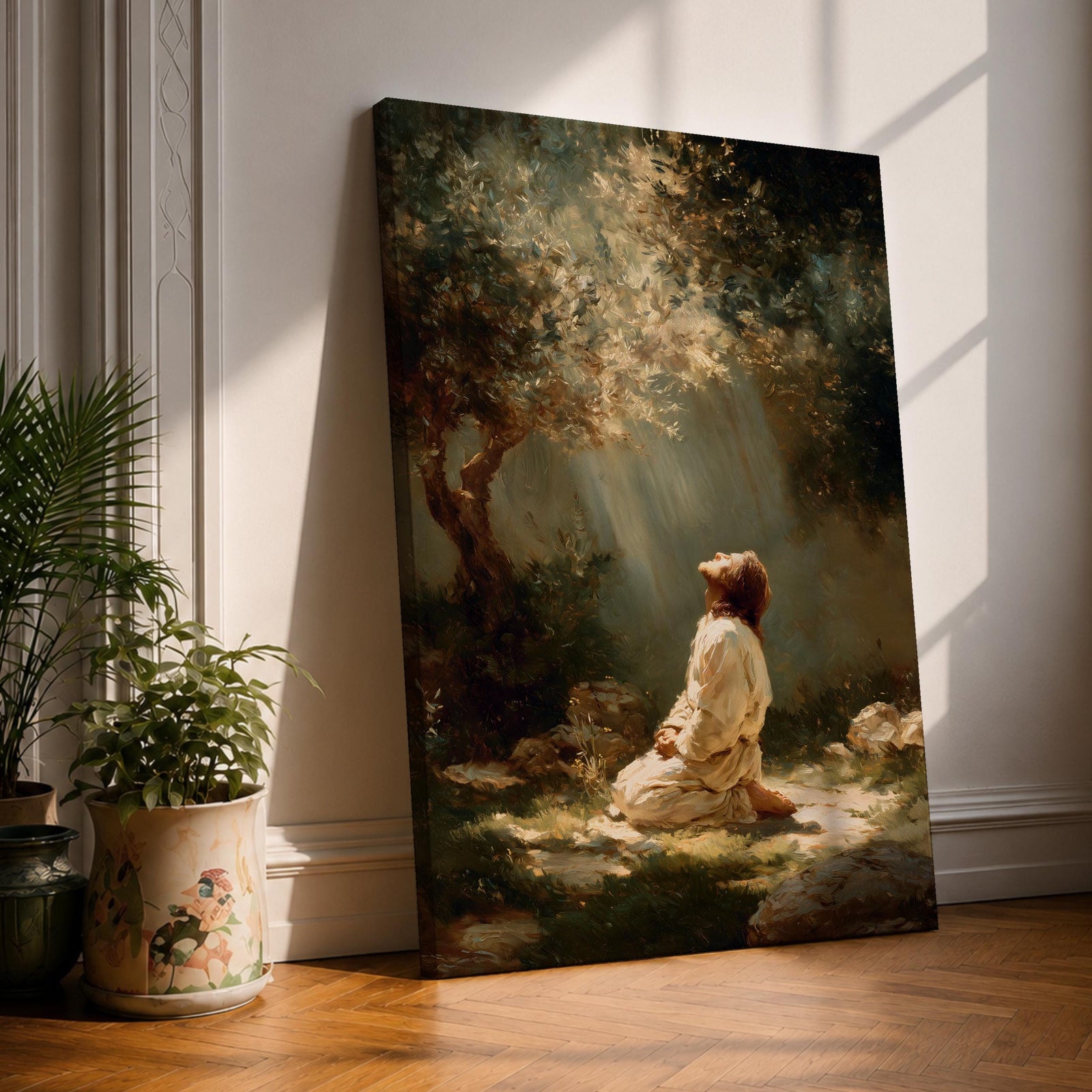 Framed artwork of Jesus praying in the Garden of Gethsemane, kneeling beneath a tree as soft rays of light shine down from above. Set in a modern living room, the canvas rests on a wooden console beside plants and neutral decor.