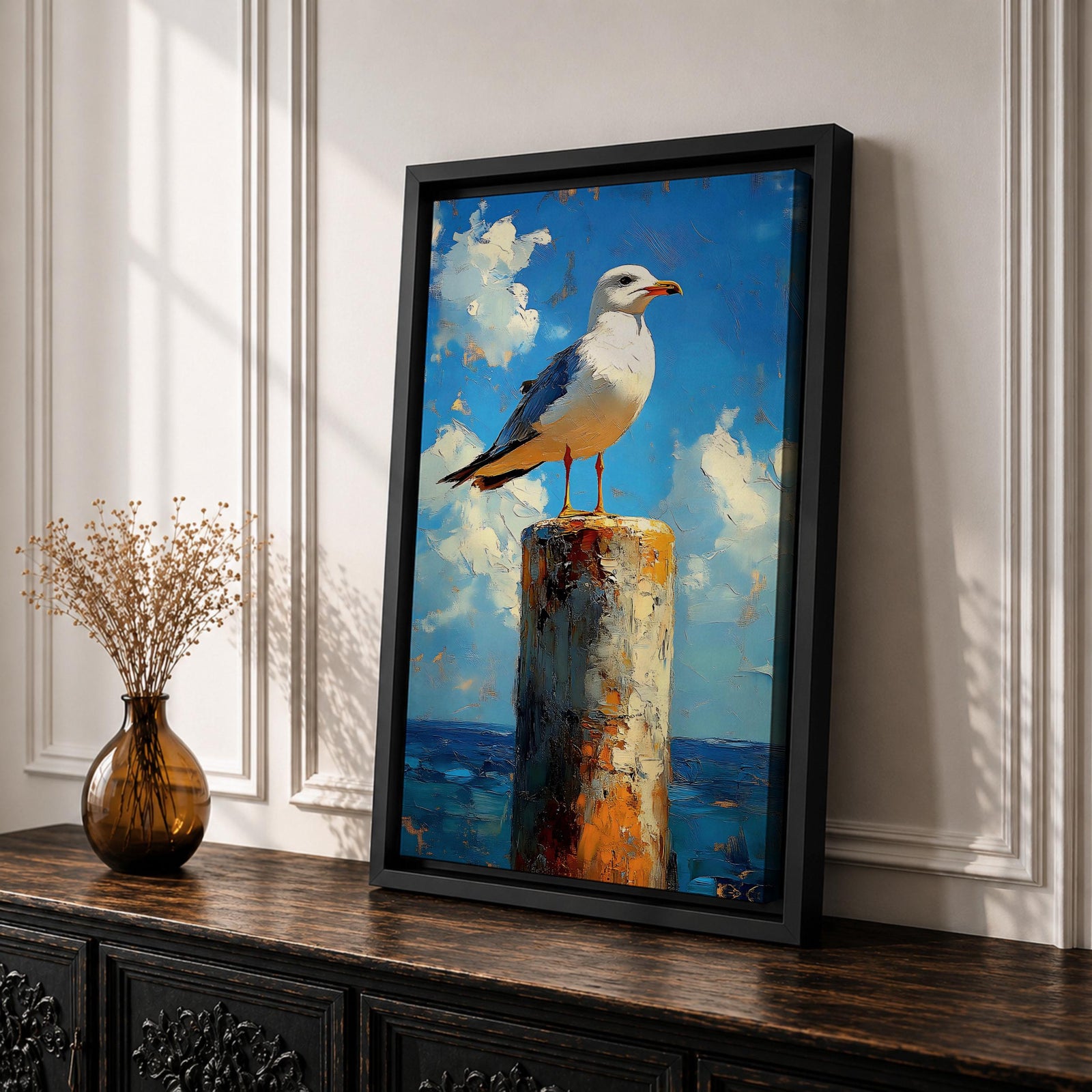 A framed canvas painting of a seagull perched on a weathered post against a bright blue sky with fluffy clouds and a distant ocean horizon. The artwork captures the bird in a realistic style with textured brushstrokes emphasizing the natural coastal scene.