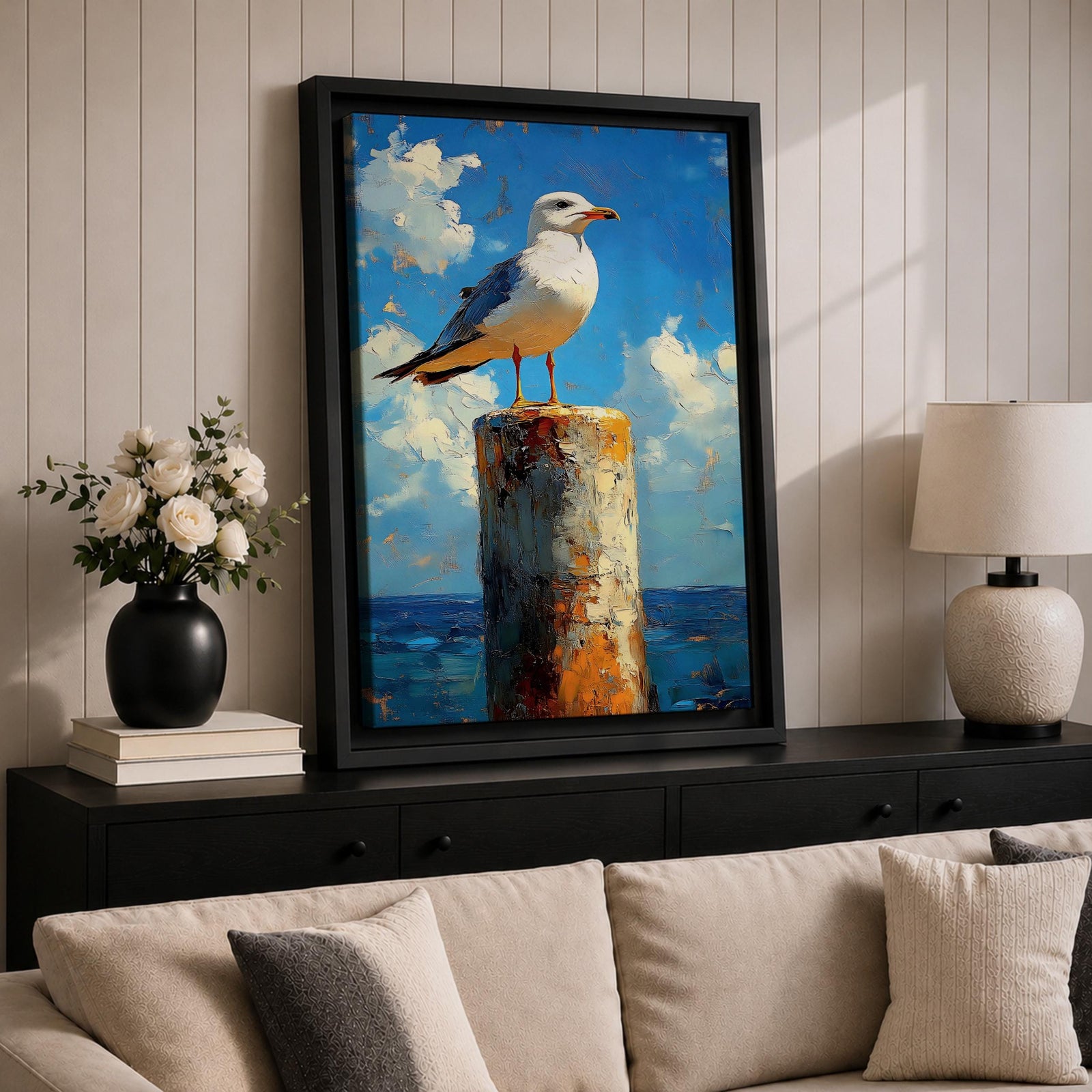 A framed canvas painting of a seagull perched on a weathered post against a bright blue sky with fluffy clouds and a distant ocean horizon. The artwork captures the bird in a realistic style with textured brushstrokes emphasizing the natural coastal scene.