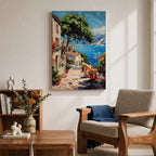 This artwork depicts a vibrant Italian village scene with terracotta-roofed houses, lush greenery, and colorful flowers lining a stone pathway that leads down to a bright blue Mediterranean sea with sailboats and distant mountains, rendered in a realistic style capturing coastal charm.
