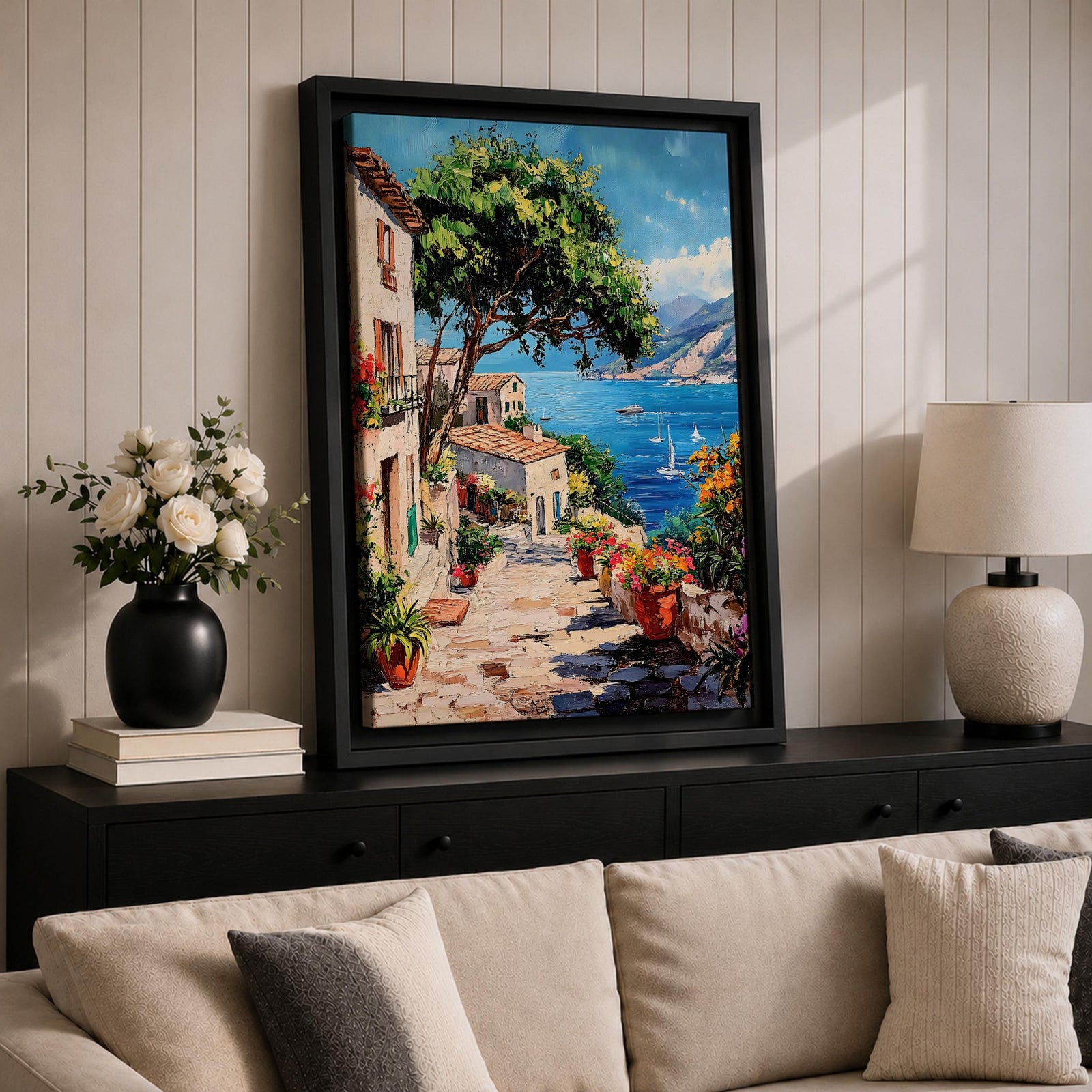 This artwork depicts a vibrant Italian village scene with terracotta-roofed houses, lush greenery, and colorful flowers lining a stone pathway that leads down to a bright blue Mediterranean sea with sailboats and distant mountains, rendered in a realistic style capturing coastal charm.