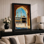 Watercolor style artwork showing the Kaaba at Masjid Al Haram in Mecca framed through a large golden mosque arch, with pilgrims gathered around the courtyard and two tall minarets rising against a clear blue sky.