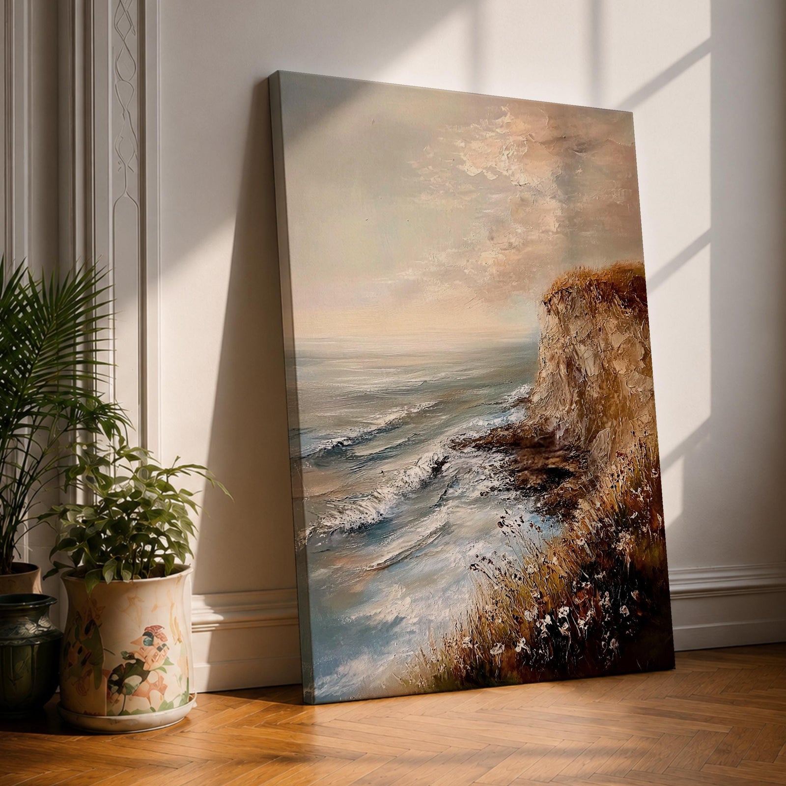 A vintage style framed canvas painting depicting a cliffside ocean seascape with waves crashing against rocky shores and wildflowers in the foreground. The textured brushwork captures the coastal landscape atmosphere with muted tones and a soft sky blending into the horizon.