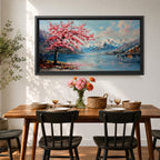 Panoramic painting of a cherry blossom tree in full bloom beside a calm mountain lake with snow covered peaks in the distance and soft clouds above. Pink petals drift across the water while thick palette knife brushstrokes create texture in the blossoms shoreline and reflections forming a wide spring landscape scene with bold color contrast.