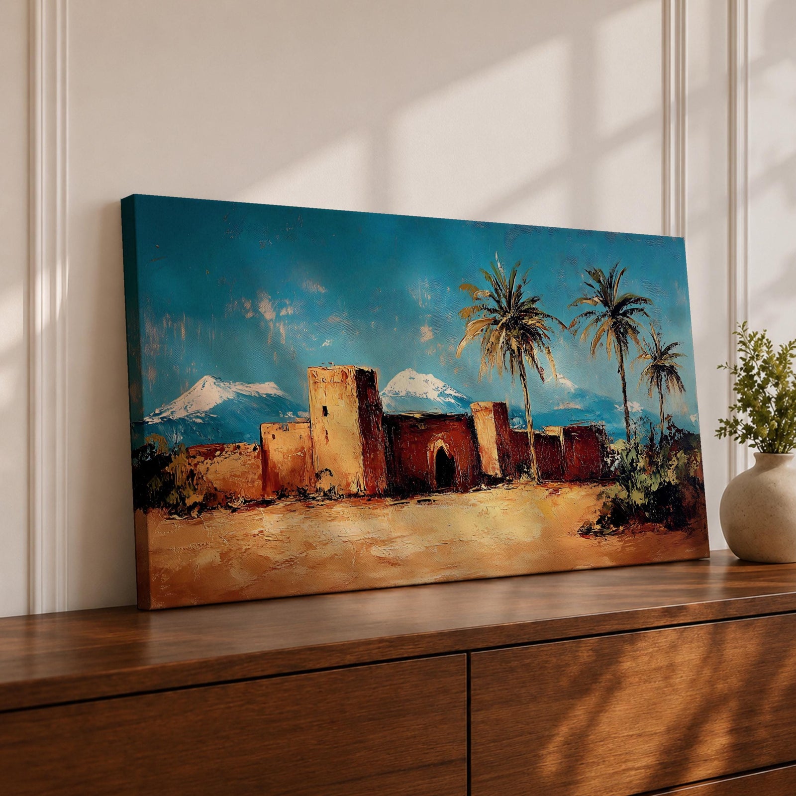 Panoramic painting of a Moroccan kasbah built from warm earthen walls set in a desert landscape with tall palm trees and distant snow capped mountains beneath a bright blue sky. Thick palette knife textures shape the fortress architecture sandy ground and foliage creating a wide horizontal North African landscape scene with bold contrast.