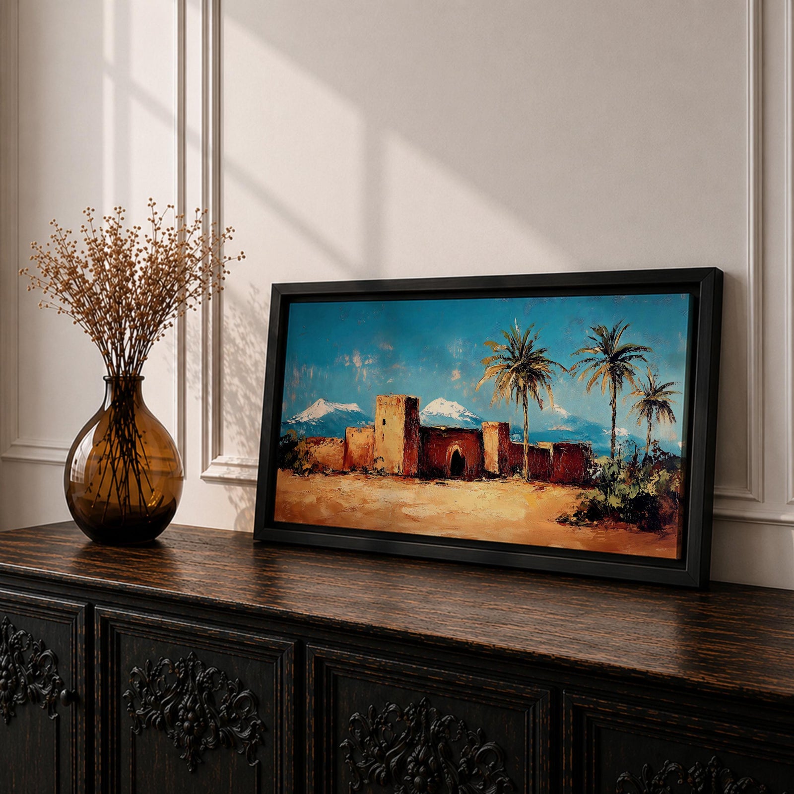 Panoramic painting of a Moroccan kasbah built from warm earthen walls set in a desert landscape with tall palm trees and distant snow capped mountains beneath a bright blue sky. Thick palette knife textures shape the fortress architecture sandy ground and foliage creating a wide horizontal North African landscape scene with bold contrast.