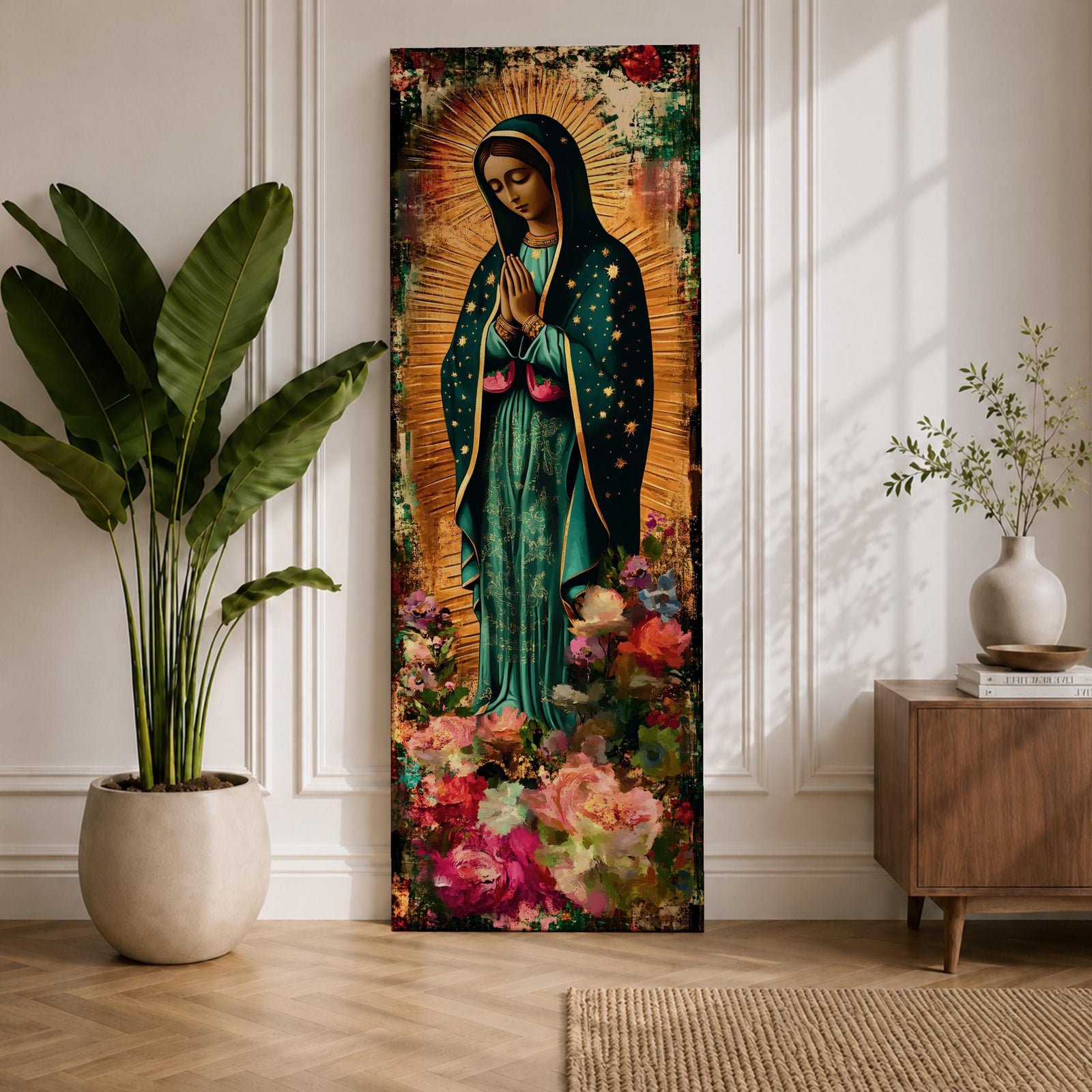 A tall narrow vertical painting of the Virgin Mary Guadalupe shown in prayer with hands clasped, wearing a deep teal mantle adorned with gold stars and a soft pink dress, surrounded by radiant golden rays, with rich floral elements in vibrant pink, red, and cream tones at the base, rendered in a textured painterly style with warm, aged tones.
