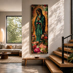 A tall narrow vertical painting of the Virgin Mary Guadalupe shown in prayer with hands clasped, wearing a deep teal mantle adorned with gold stars and a soft pink dress, surrounded by radiant golden rays, with rich floral elements in vibrant pink, red, and cream tones at the base, rendered in a textured painterly style with warm, aged tones.