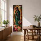 A tall narrow vertical painting of the Virgin Mary Guadalupe shown in prayer with hands clasped, wearing a deep teal mantle adorned with gold stars and a soft pink dress, surrounded by radiant golden rays, with rich floral elements in vibrant pink, red, and cream tones at the base, rendered in a textured painterly style with warm, aged tones.