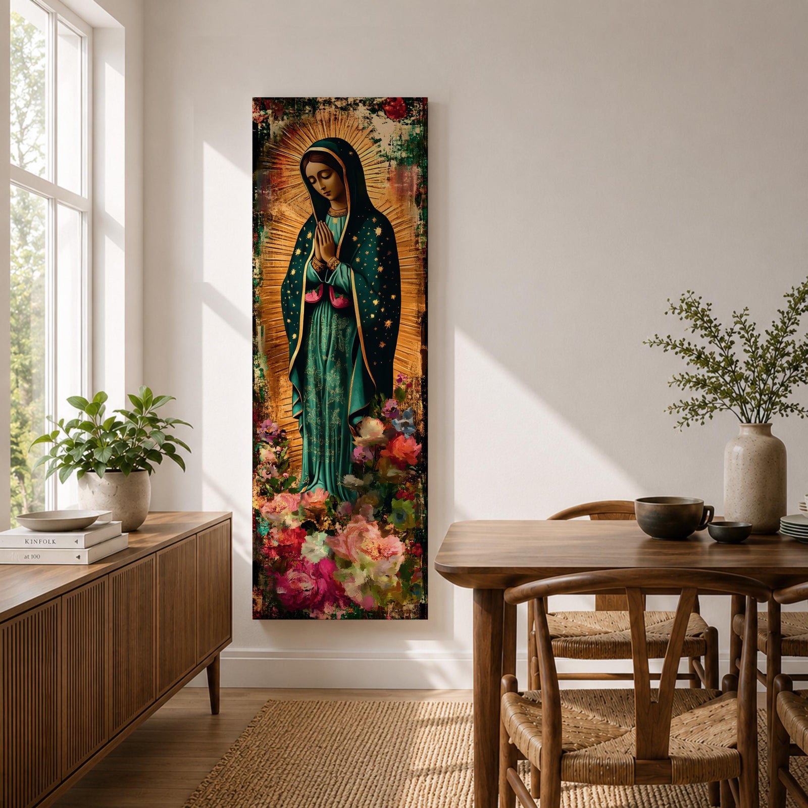 A tall narrow vertical painting of the Virgin Mary Guadalupe shown in prayer with hands clasped, wearing a deep teal mantle adorned with gold stars and a soft pink dress, surrounded by radiant golden rays, with rich floral elements in vibrant pink, red, and cream tones at the base, rendered in a textured painterly style with warm, aged tones.
