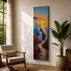 A tall vertical painting depicts a Santorini coastal scene with whitewashed buildings perched along a cliffside, a winding stone path leading toward the sea, and a glowing sunset casting warm orange and gold light across the sky and water, with a windmill silhouette and small sailboats adding depth to the vibrant Mediterranean composition.