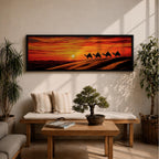 Panoramic desert sunset painting featuring a silhouette caravan of camels and riders walking along rolling sand dunes beneath a glowing red and orange sky. The low sun sits near the horizon while layered desert dunes stretch into the distance, creating a dramatic wide composition across the desert landscape.
