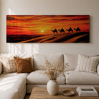 Panoramic desert sunset painting featuring a silhouette caravan of camels and riders walking along rolling sand dunes beneath a glowing red and orange sky. The low sun sits near the horizon while layered desert dunes stretch into the distance, creating a dramatic wide composition across the desert landscape.