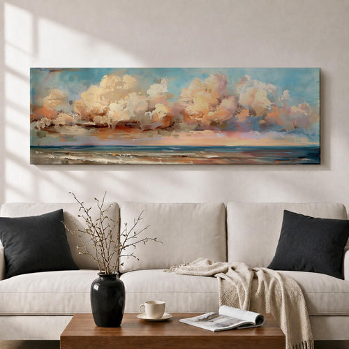 Panoramic coastal seascape painting of ocean waves reaching a sandy shoreline beneath expansive billowing clouds glowing with warm sunset tones. Soft peach, cream, and blue hues blend across the sky while gentle reflections shimmer along the water in a wide horizontal seaside composition.