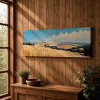 Panoramic wheat field landscape painting showing rolling golden farmland stretching across hills beneath a bright blue sky with scattered clouds. Textured brushstrokes depict tall wheat grasses in the foreground leading toward layered countryside fields, tree clusters, and distant hills across the wide rural horizon.