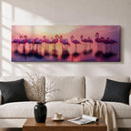 Panoramic artwork showing a flock of flamingos standing in shallow reflective water at sunset. The long horizontal composition features pink birds mirrored in the calm lake surface with glowing orange, purple, and magenta sky tones behind them, creating a tropical waterscape with silhouettes and soft reflections stretching across the scene.