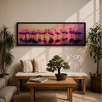 Panoramic artwork showing a flock of flamingos standing in shallow reflective water at sunset. The long horizontal composition features pink birds mirrored in the calm lake surface with glowing orange, purple, and magenta sky tones behind them, creating a tropical waterscape with silhouettes and soft reflections stretching across the scene.
