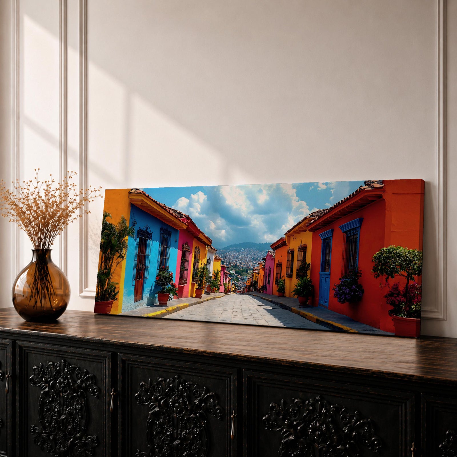 Panoramic cityscape painting showing a vibrant Mexican street lined with colorful colonial houses in bright shades of blue, red, orange, and yellow, with potted plants and balconies along a cobblestone road leading toward distant hills beneath a partly cloudy sky.