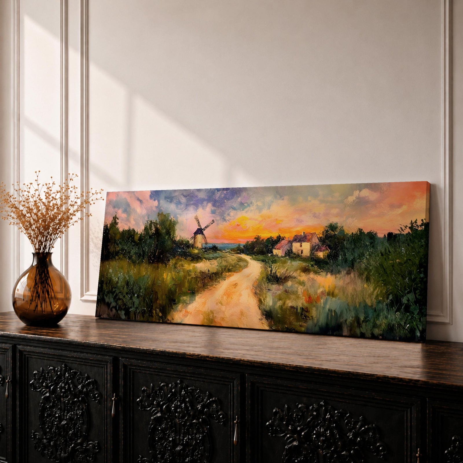 A panoramic countryside landscape painting showing a winding dirt road leading through grassy fields toward a traditional windmill and small farmhouse under a glowing sunset sky, with soft clouds and warm golden light illuminating the rural scenery across the wide composition.